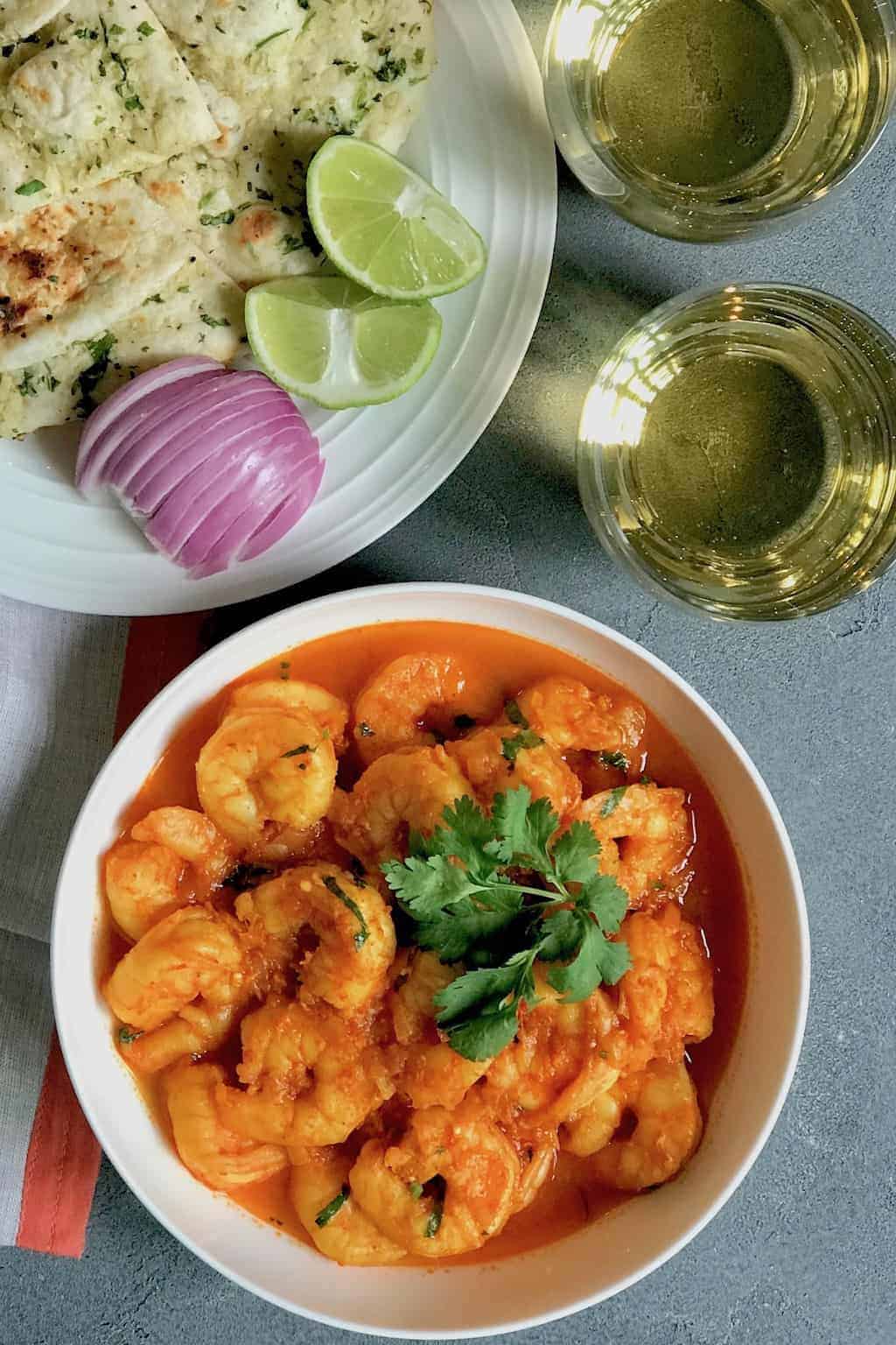 Shrimp curry in a white serving bowl, a white plate with cut up naan, sliced red onions and lemon wedges. Sparking drink in 2 cups on the side.