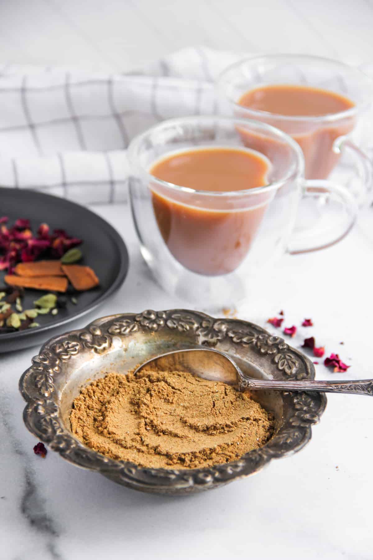 Chai masala powder in a bowl along side whole spices and masala chai 