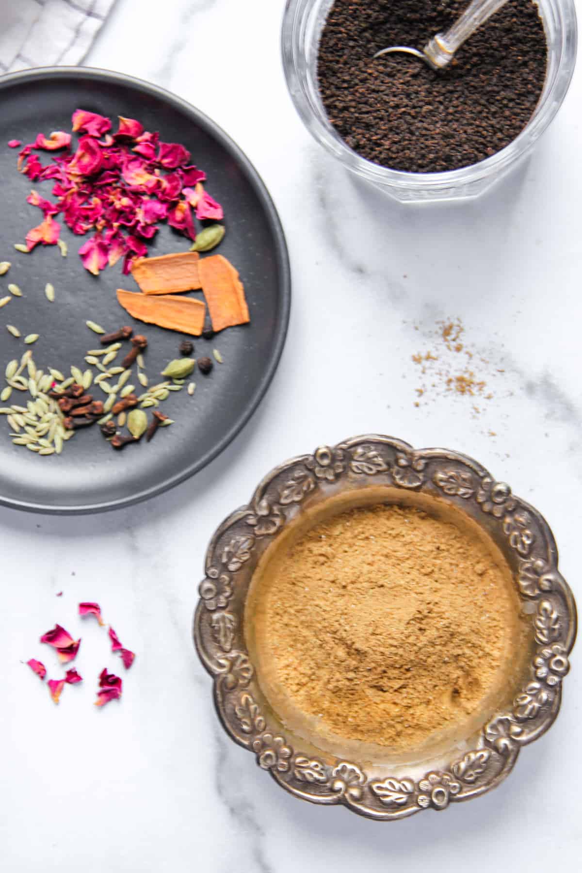 Chai masala powder in a bowl along side whole spices and loose black tea 