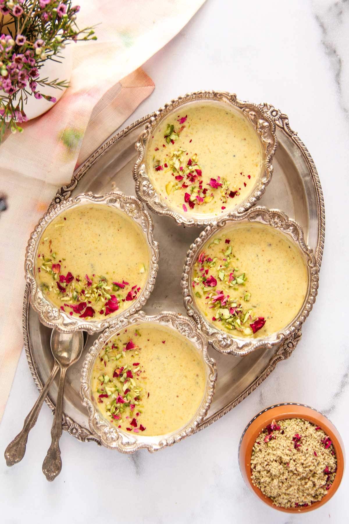 Thandai Phirni served in silver bowls on a silver tray garnished with pistachios and rose petals