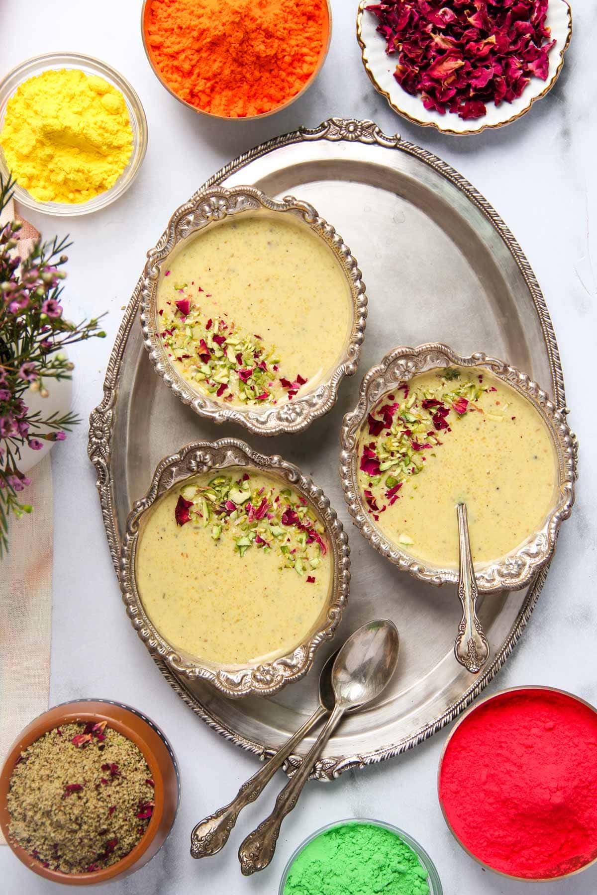 Thandai Phirni served in silver bowls garnished with pistachios and rose petals with bowls of holi colors on the sides